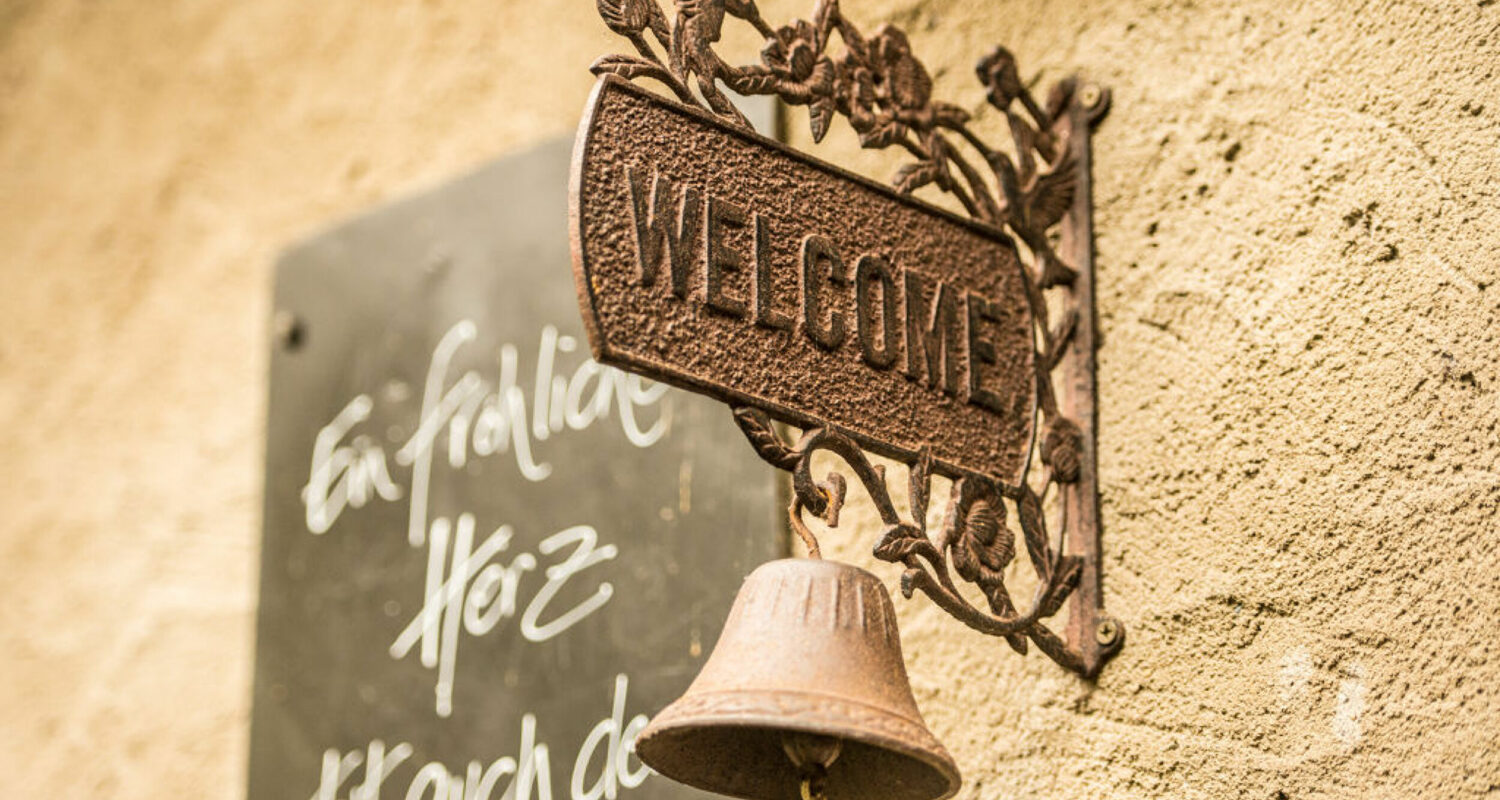 Dekoratives Bild: Rostfarbenes Metallschild mit der Aufschrift „WELCOME“ und einer kleinen Glocke hängt an einer verputzten Außenwand; daneben steht ein handgeschriebener Text auf einer Tafel.