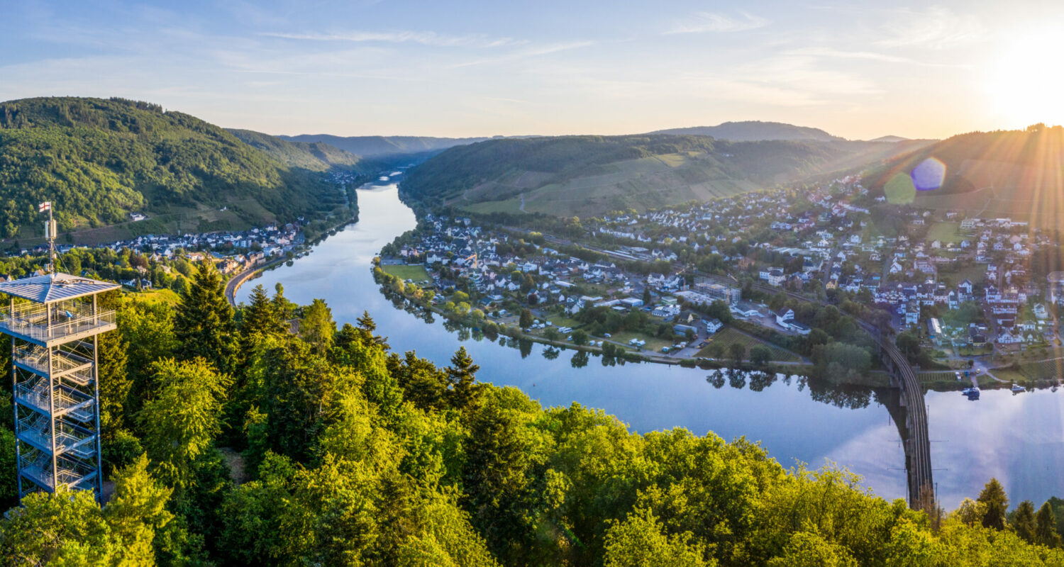 Dekoratives Bild: Luftaufnahme mit Blick auf den Aussichtsturm Prinzenkopf an der Mosel. Die Mosel fließt im tiefstehenden Sonnenlicht durch dicht bewaldeten Hänge und an einer Ortschaft entlang.