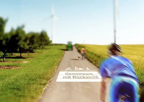 bodenmarkierung ©️ Rheinhessen Touristik Radweg durch eine ländliche Landschaft mit Radfahrenden und dem Hinweis auf dem Weg „Gemeinsam mit Rücksicht“.