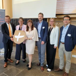 Parlamentarisches Frühstück Sechs Personen, darunter Staatssekretärin Petra Dick-Walther, stehen in einem Raum und halten ein goldenes rundes Schild mit einem weißen Plus-Zeichen.