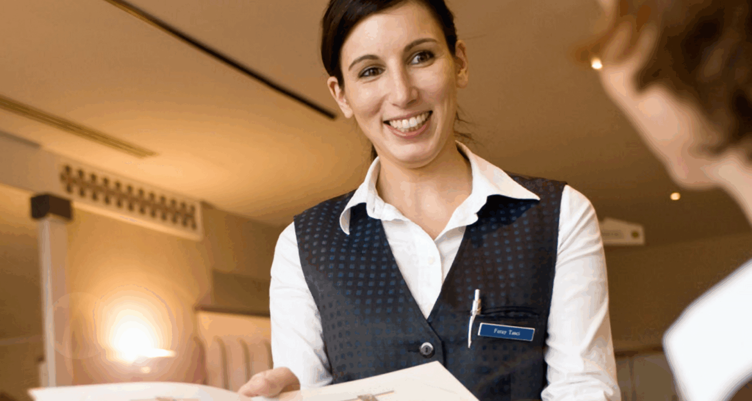 Eine Servicemitarbeiterin in einem Hotel erklärt einem Gast etwas. In der Hand hält sie einen Plan