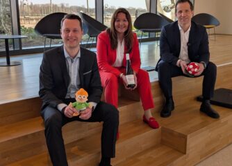 Pressegespräch zum Tourismusjahr 2025 in Mainz Drei Personen sitzen auf einer Holzstufe in einem modernen Gebäude mit großen Fenstern. Sie tragen Businesskleidung und halten symbolische Gegenstände in den Händen, darunter eine Weinflasche, einen Fußball und eine kleine Figur. Hinter ihnen ist eine Glasfront mit Blick nach draußen zu sehen.